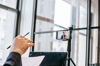Fotografia de uma pessoa em um ambiente de escritório, participando de uma videoconferência ou gravação usando um smartphone montado em um tripé. A câmera do smartphone está voltada para a pessoa, que aparece na tela do dispositivo enquanto fala ou apresenta algo. A mão de outra pessoa, segurando uma caneta e um pedaço de papel, também aparece em primeiro plano, sugerindo que a pessoa está envolvida na conferência, talvez como espectadora ou participante