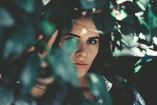 White woman with brown eyes and hair standing in the shade. Her face is half covered by leaves hanging from a tree. Only one of her eyes is shown. Through the blurry leaves you can see she’s holding up her hand and pushing leaves out of her face. It seems as though the photo was made on a sunny day.