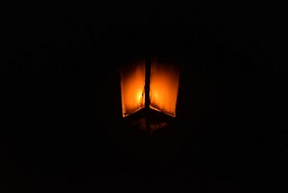 A canted view from below of what looks like a gas streetlamp. It glows orange is enveloped in a completely black background.