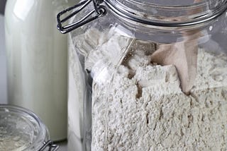 Close up of a jar with white powder and a scoop in it against a background of a bottle with milk