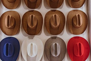16 cowboy hats hanging on a wall, they are all the same color except for the third row where they are blue, white, silver and red, while all the others are brown