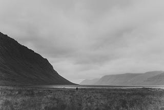 Black and White photo of a person standing in a valley plateau with rising cliffs on one side.