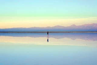 Single figure standing on water which is reflecting a clear sky