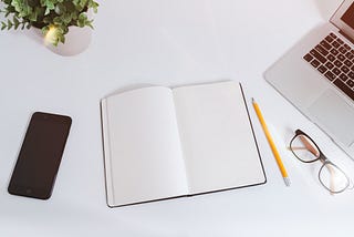 A phone, notebook, pencil, glasses, plant, and laptop on a table.