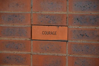 A picture of an orange-brown brick wall, with the center brick written ‘COURAGE’