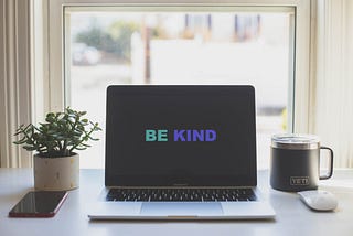 A desk with a laptop in the middle with the words “BE KIND” on the screen.