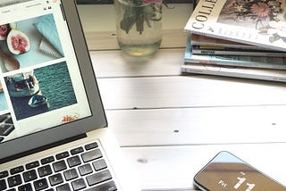 A workdesk with a MacBook, mobile phone, books and a vase.