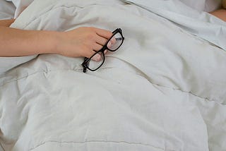 A lady sleeping tucked in white covers, eyeglass in hand, pillow partly above face.