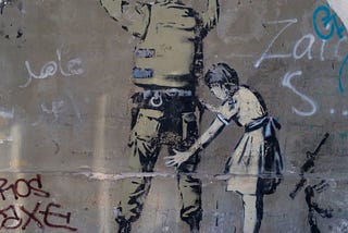 Graffito on a wall, showing a soldier with his back turned towards the viewer and his hands in the air. A little girl is performing a body search on the soldier, making the picture a subversion of usual expectations.