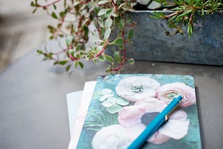 A floral-covered journal and pen, and a metal plant holder and plant on an outdoor table