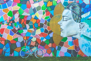 A teal bicycle is parked in front of a multicolored mosaic mural with a woman’s facial profile.