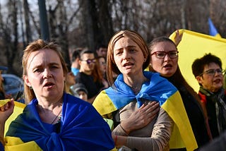 Two women draped in Ukraine’s yellow and blue flag sing while standing outside among a crowd of others protesting the war in Ukraine. One women raises her fist and the other woman holds her right hand over her heart.
