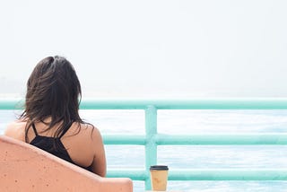 A woman from behind, who has dark hair down to her shoulders, and is wearing a black halterneck vest top. She is sitting in a pink seat with a takeaway coffee next to her and she is looking out at the sea. There is an aqua-blue divide between her and the sea, like she is on a boat.