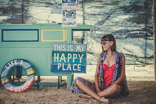 Lady smiling as she sits next to a sign which reads This is my Happy Place.