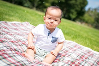 Asian baby sitting on a plaid blanket over grass. He is ostensibly frowning.