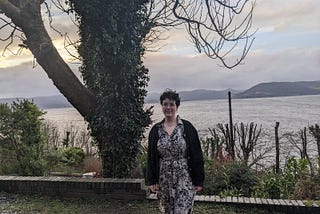 Kirstie standing in a landscape, with an ivy-covered tree, a body of water, and a distant shore all visible. The sky has patches of blue and a mix of bright and grey clouds.
