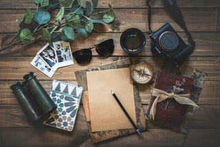 A desk with a notebook, pencil, binoculars, photos, sunglasses, a cup of coffee, a small branch, a compass, a camera and a journal sitting on top.