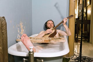 A video still of Ragnar Kjartansson in a full bathtub playing the guitar and wearing headphones.