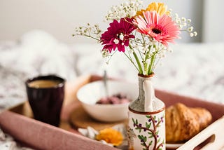 Breakfast tray on bed with coffee, pastry, yogurt, and flowers.