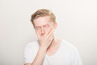 The image shows a person with light skin and reddish-blonde hair, wearing a white shirt. The person has their eyes closed and their hand covering most of their face, as if they are expressing frustration, exhaustion, or guilt. The background is plain and light-colored, creating a neutral setting that focuses on the individual’s expression and gesture.