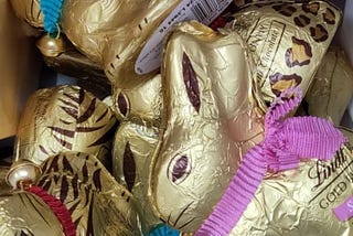 Close-up of a jumble of Lindt chocolate bunnies. They are covered in gold foil with rabbit eyes, ears, and whiskers printed in brown. Each bunny wears a coloured ribbon as a collar with a little bell on it
