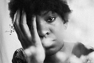 A black and white portrait of a Black woman with a short afro. Her hand covers her eye. White flowers sit in her hair and partially cover her face. She is wearing a floral dress.