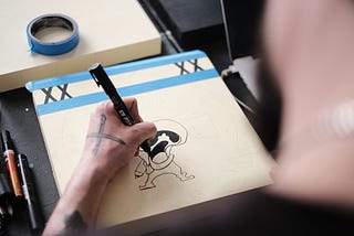 An illustrator tightly grips an ink pen as they draw a cartoon on a sheet of high quality expensive paper, which is held to a board with masking tape. His tools and materials and scattered around.