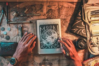 Human hands place on a table covered by a map, other materials and a book of the moon.