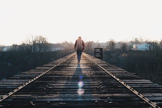 A man with a backpack walks alone down the middle of train tracks