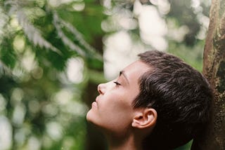 Woman leaning against a tree with her eyes closed, green leaves and sunlight in the background.