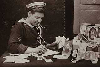 Sepia-tone old-fashioned photo of a sailor smiling while writing a letter with a fountain pen and inkwell on stationery — family photos sit framed on the desk beside him.