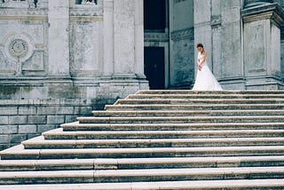 A woman in a wedding dress standing alone at the top of stairs at a church. Her head is down and her body language says she is disappointed and sad. This signifies the unmet expectations we can experience during separation on the Twin Flame journey.