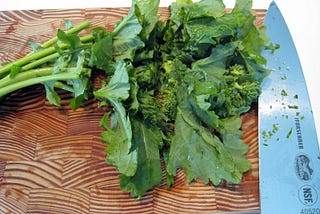 A bunch of broccoli rabe on a cutting board with a chef’s knife