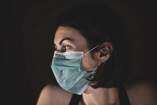 A woman with black chin length hair tucked beind her ear, is wearing a face mask and facing the right. She is wearing a black singlet and standing in front of a black background.