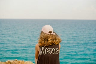 A person with long hair and a baseball cap looks out to sea with ‘misfits’ written on the back of their sleeveless t-shirt.