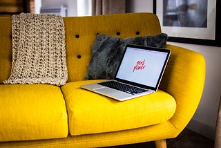 A colour photo of an open laptop on a yellow couch with a blanket and a cushion.