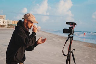 a guy on a beach standing in front of a camera looking into the camera with his hands towards his face