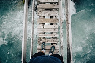 The view of the picture is a person looking down at his feet over a wooden bridge with a river underneath him.