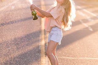 A happy girl popping a bottle of champagne in the middle of a tree-lined street