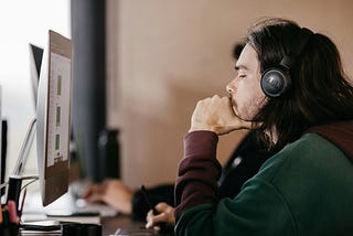 A man with long hair is sitting in front of a computer screen. He is wearing a headset, and his expression conveys deep thinking.