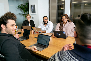A group of people sit around a table, in a conference room. In front of each person is a laptop and some of the laptops have stickers placed on the back.