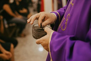 A robed priest holding a glass vessel containing ashes for Ash Wednesday. The view is from the side. The priest’s hands and the vessel are central, a small portion of their torso is visible, and the parishioners on the lefthand side of the image are blurred.