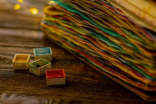Stack of different colored papers with four small paint containers in front of yellow, green, blue, and red paint.