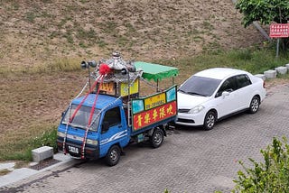 Looking down at a road with two parked vehicles. One is an ordinary white car. The other is a small blue cabbed truck with a traditional Chinese structure on top, with loudhailers either side pointing front and back. Behind the vehicles, a dry, scrubby grass bank slopes upwards