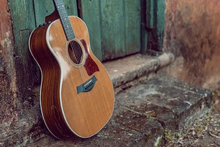 A tan-wooded guitar standing on a weathered stoop next to a green-painted barn-like door.