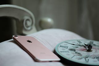 Corner of white bed with pink iphone 8 and alarm clock face lay across it