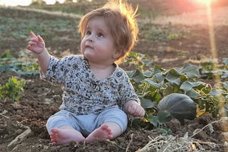 A barefoot baby sitting beside a melon in a field, a sun rising behind them, points upwards.