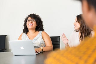 Fun at work. Conference room. One woman types on her MacBook, laughing as her coworker tells an animated story.