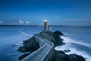 A lighthouse at the end of a long walkway begins to shine at dusk.