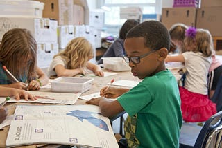 A group of young children are seated at a classroom table, focused on drawing and coloring in their workbooks.